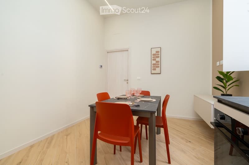 A modern dining area with a sleek black dining table, six orange chairs, and a small potted plant on the counter. The room features light wooden flooring and white walls.