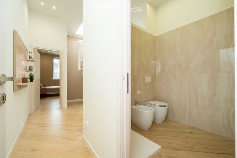 A modern hallway with light wooden flooring, white walls, and a small shelf with decorative items. The hallway leads to a bathroom with a toilet and bidet, and a bedroom with a bed and window.