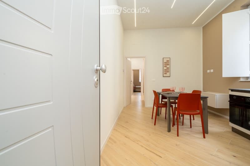 Modern hallway leading to a dining area with a grey dining table, red chairs, and a built-in oven.