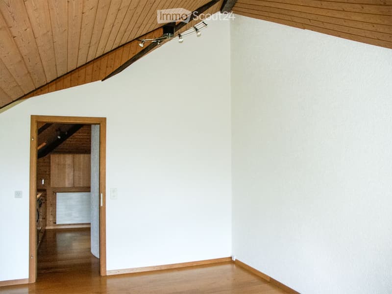 Empty room with wooden flooring, white walls, and a wooden ceiling with track lighting. An open doorway leads to another room with cabinets and a staircase.
