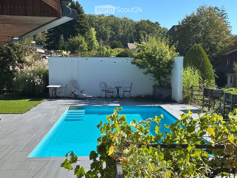 A well-maintained garden with a swimming pool, surrounded by greenery, outdoor furniture, and a white wall. The area includes a patio with tables and chairs, and a wooden fence.