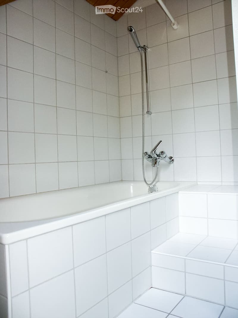 Clean, modern bathroom with white tiled walls and floor, featuring a bathtub with a modern faucet and a handheld showerhead.
