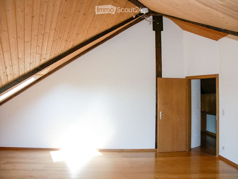 Empty attic room with wooden slanted ceiling, white walls, wooden floor, and a single wooden door leading to another space.
