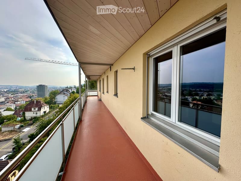 Balcony with wooden ceiling, red floor, metal railing, large windows offering a cityscape view.