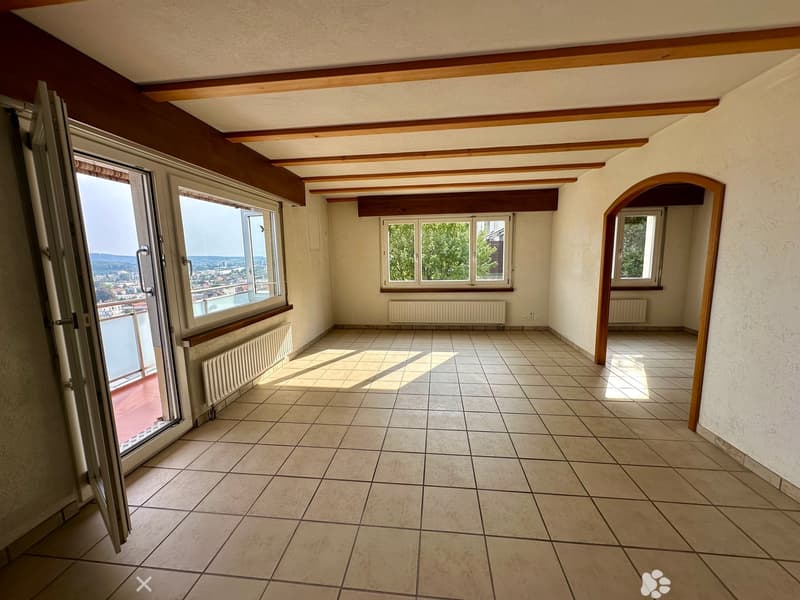 Empty room with tiled floor, large windows, and wooden beams on the ceiling. Radiators are visible along the walls.