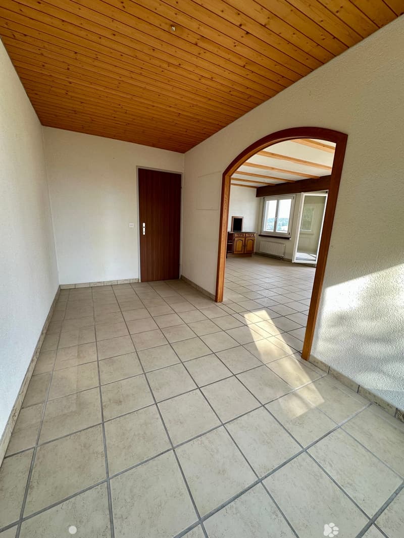 Empty hallway with tiled floor, wooden ceiling, and an arched doorway leading to another room with a TV and cabinet.