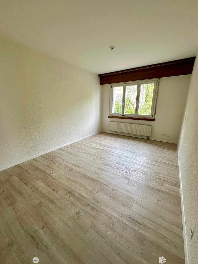 Empty room with light wooden flooring, white walls, and a window with white frames. A radiator is placed below the window.