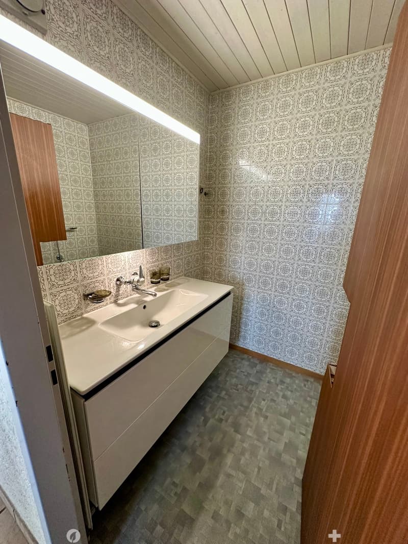 Modern bathroom with a white sink, chrome faucet, and a large mirror above it. The walls are tiled with a decorative pattern, and the floor has a patterned tile design.
