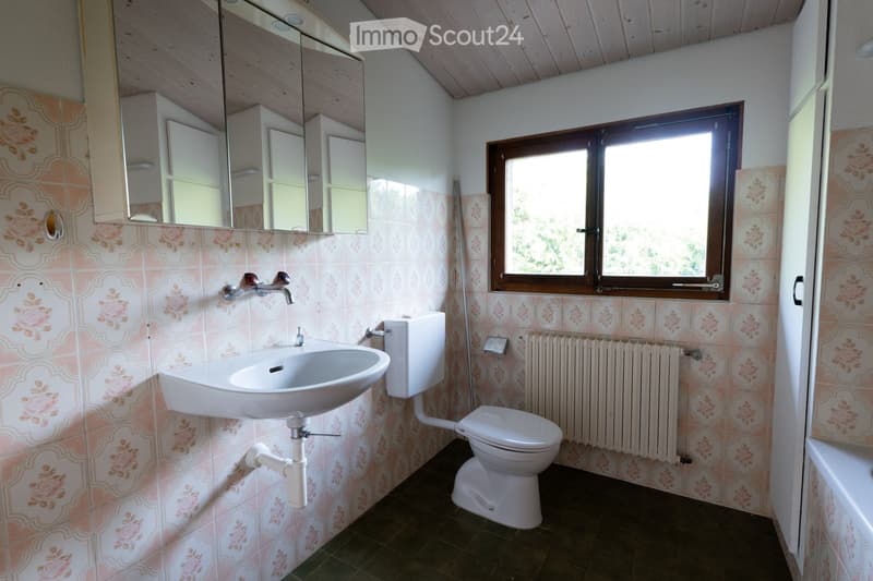 Bathroom with pink floral tiles, a white sink, toilet, mirror, and a window with wooden frame. Radiator is visible below the window.