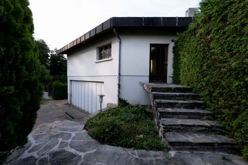 A modern white house with a flat roof, garage, and ivy-covered wall, situated on a stone pathway with steps leading to the entrance.