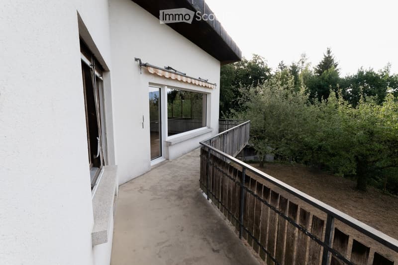 Balcony with a wooden railing, concrete floor, and a view of a garden with trees and shrubs.