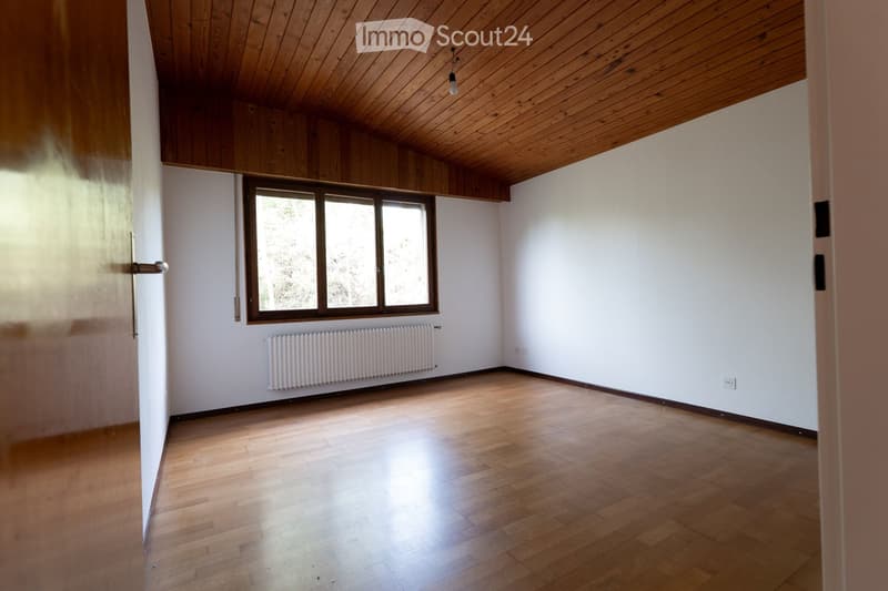 Empty room with wooden flooring, white walls, large window, radiator, and wooden ceiling beams.