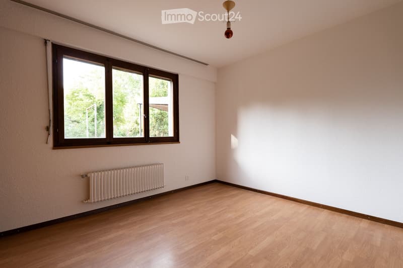 Empty room with light-colored walls, wooden flooring, large window with brown frame, radiator, and a single hanging light fixture.
