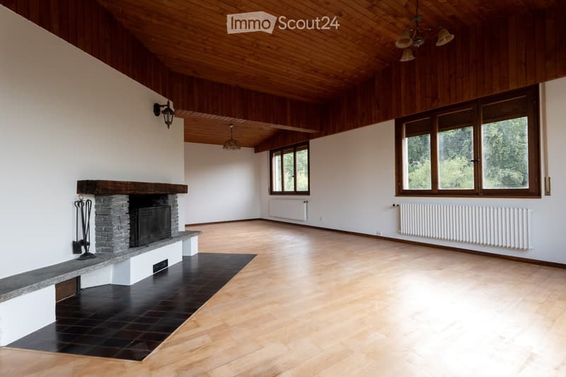 Spacious, unfurnished room with wooden floors, white walls, large windows, and a stone fireplace. The room features exposed wooden beams on the ceiling and radiators along the walls.