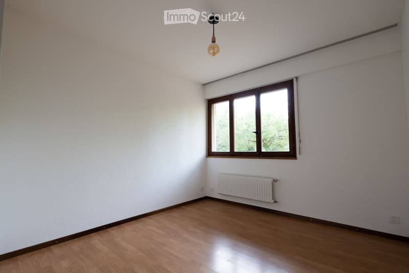Empty room with wooden flooring, white walls, a window with wooden frame, a radiator, and a single hanging light fixture.