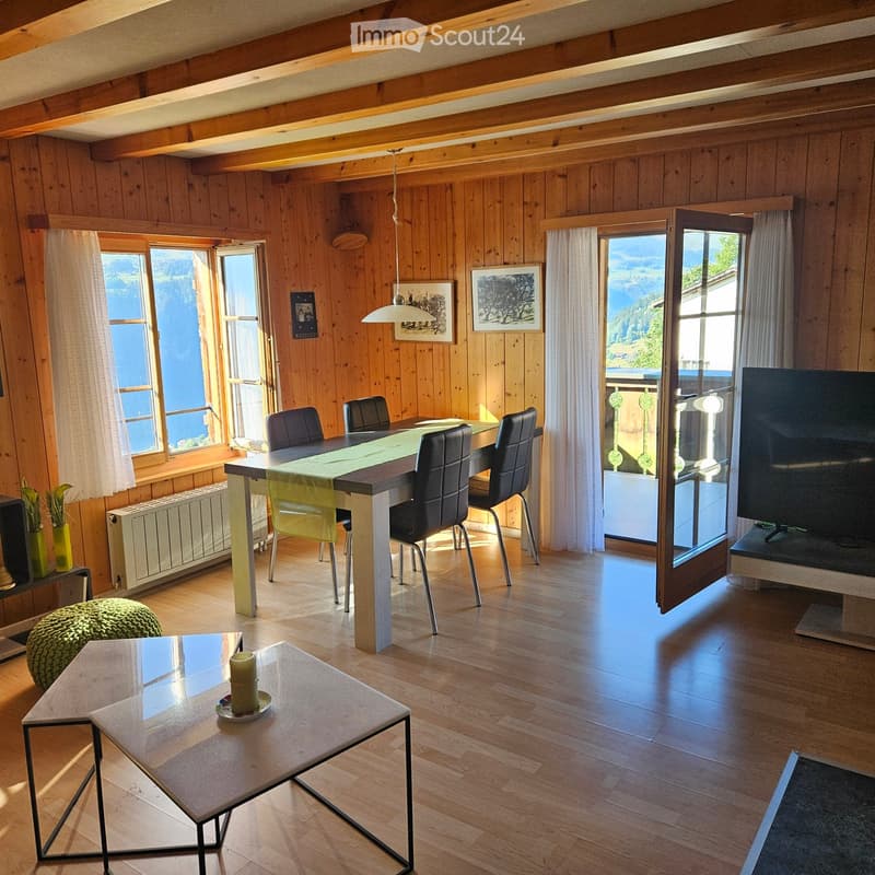Cozy living and dining area with wooden walls and floors, large windows offering a scenic view, a dining table with chairs, a glass coffee table with a candle, a green stool, and a television on a stand.