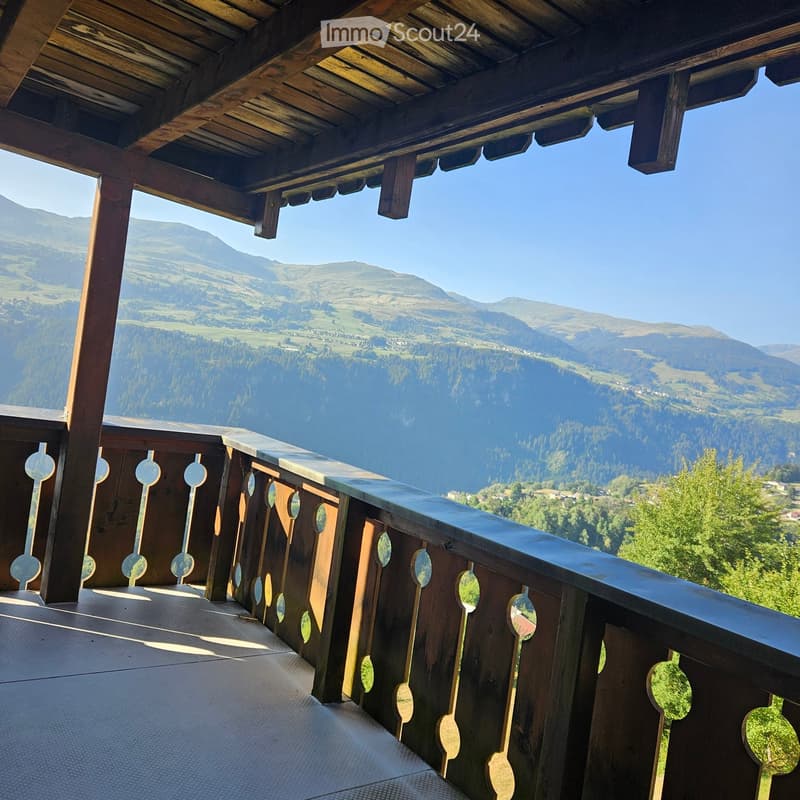 Wooden balcony with a railing featuring circular cutouts, overlooking a scenic mountain landscape with valleys and forests.
