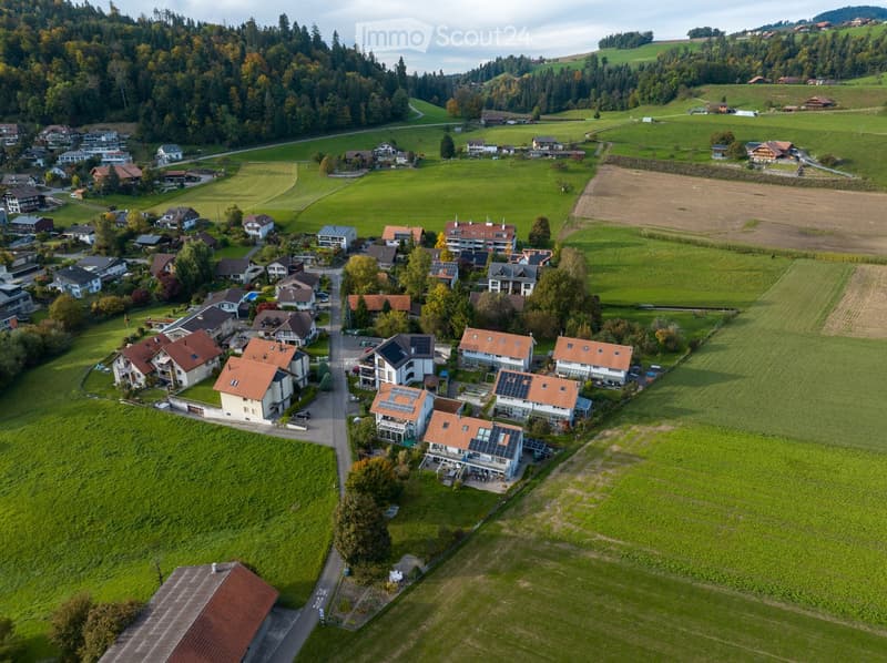 Aerial view of a residential area with multiple houses, surrounded by green fields and forested hills.