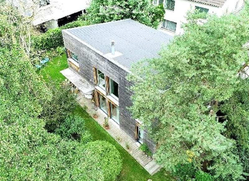 A modern, single-story house with a dark grey facade and large windows, surrounded by greenery and trees.