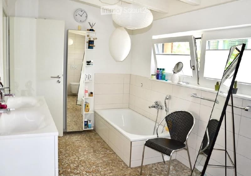 Bright bathroom with a bathtub, a black wicker chair, a large mirror, a sink, and a shelf with toiletries. The room features white tiles and a decorative pendant light.