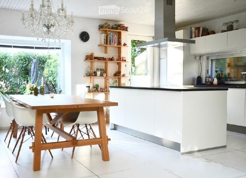 Bright, modern kitchen with white cabinets, a central island, a wooden dining table with chairs, shelves with jars and books, and a chandelier.