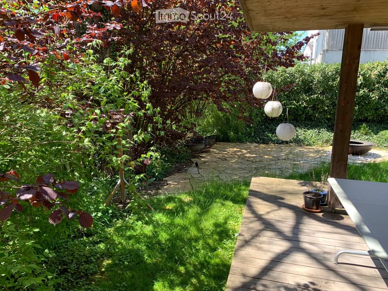Garden with wooden deck, hanging lanterns, potted plants, and lush greenery.