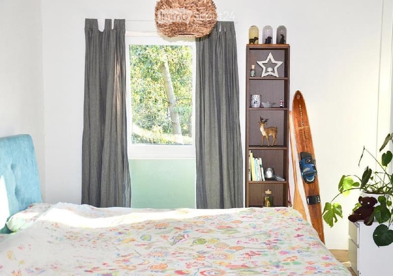 A cozy bedroom with a bed, a large window with grey curtains, a wooden shelf with books and decorative items, a snowboard leaning against the wall, and a potted plant near the window.