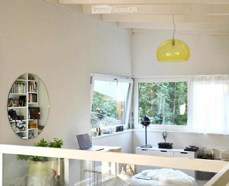 Bright, modern room with large windows offering a view of greenery, featuring a laptop on a table, a bookshelf with books and decorative items, a round mirror, and a small ottoman with a white fur throw.