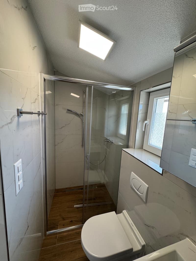 Modern bathroom with a toilet, sink, glass-enclosed shower, and a large window providing natural light. The walls are tiled in a marble pattern, and the floor is wooden.