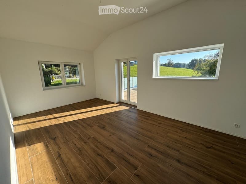 Empty room with wooden flooring, white walls, and large windows offering a view of greenery outside. Features include a sliding glass door leading to a wooden deck.