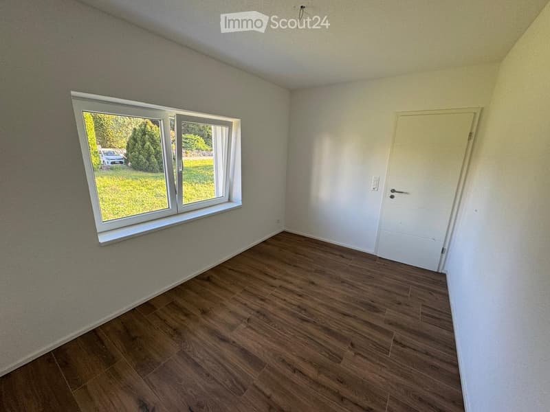 Empty room with wooden flooring, white walls, large window with view of greenery, and a white door.