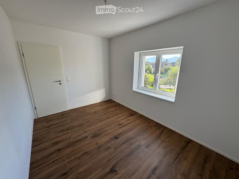 Empty room with wooden flooring, white walls, a white door, and a window showing a view of greenery and mountains.