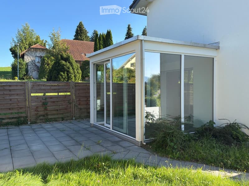 Modern glass-enclosed patio attached to a house, with a wooden fence, paved patio, and surrounding greenery.