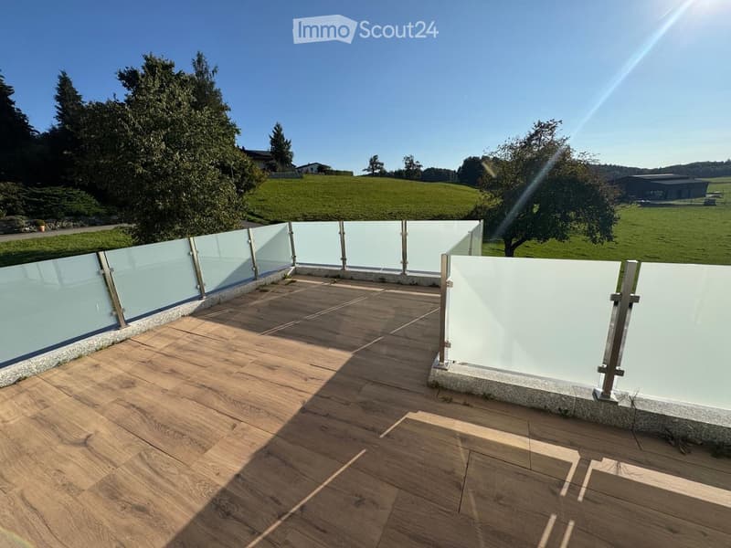 Wooden deck with glass railings, overlooking a green field and trees under a clear blue sky.