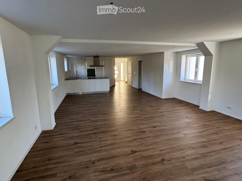 Empty room with wooden flooring, white walls, and large windows. The room is connected to a small kitchen area with white cabinets and a sink.