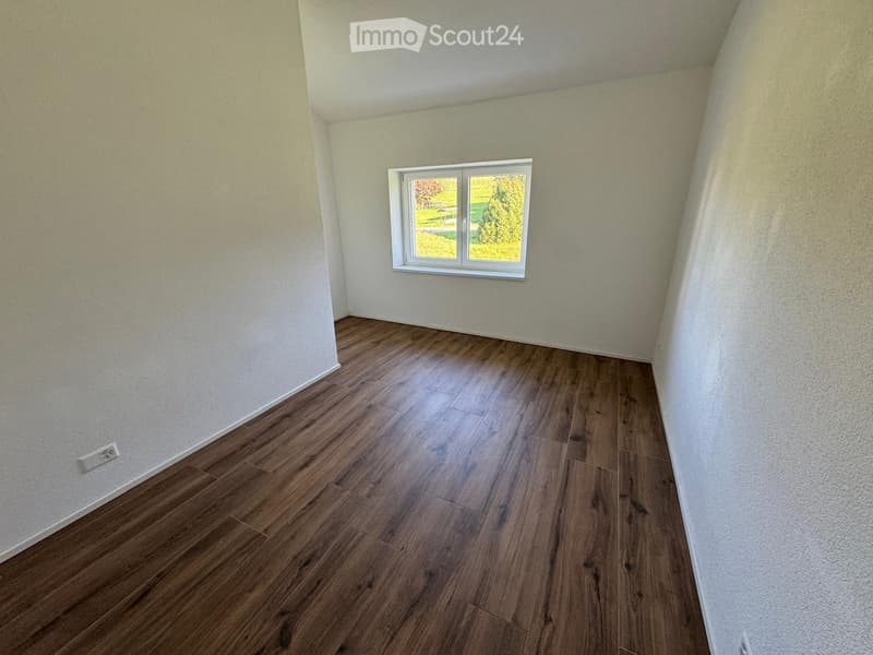 Empty room with wooden flooring and white walls, featuring a single window with white frame and a visible electrical outlet on the left wall.