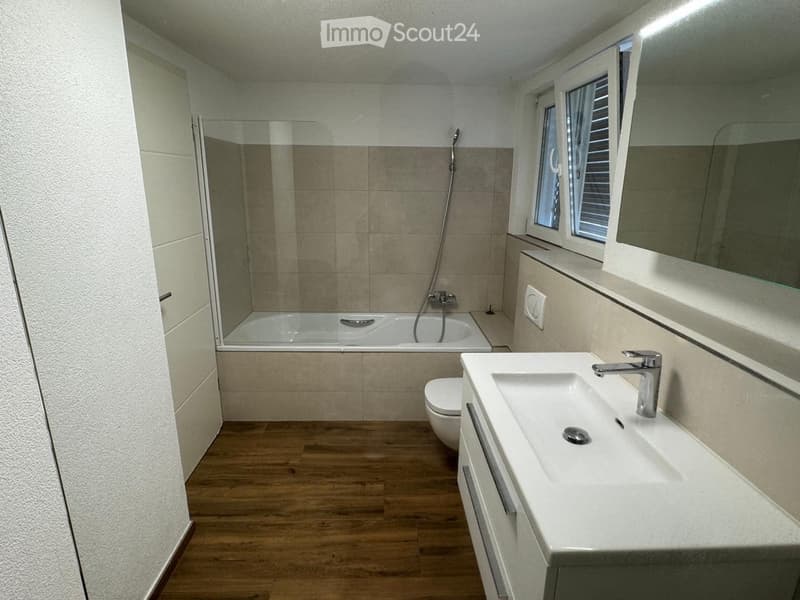 Modern bathroom with a bathtub, sink, toilet, and large mirror. Features wooden flooring and a glass-enclosed shower area.