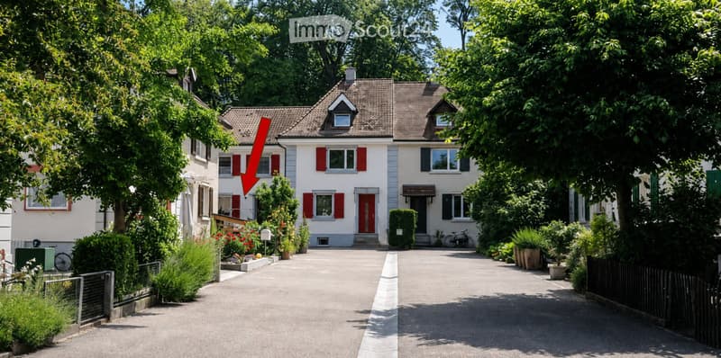 Two-story house with red shutters, white walls, and a tiled roof. The house features a driveway, garden, and is surrounded by trees and greenery.