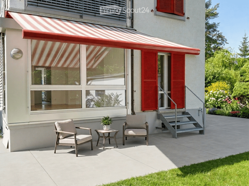 Outdoor patio area with two chairs, a small table with a potted plant, and a staircase leading up to a building entrance with red shutters and a striped awning.