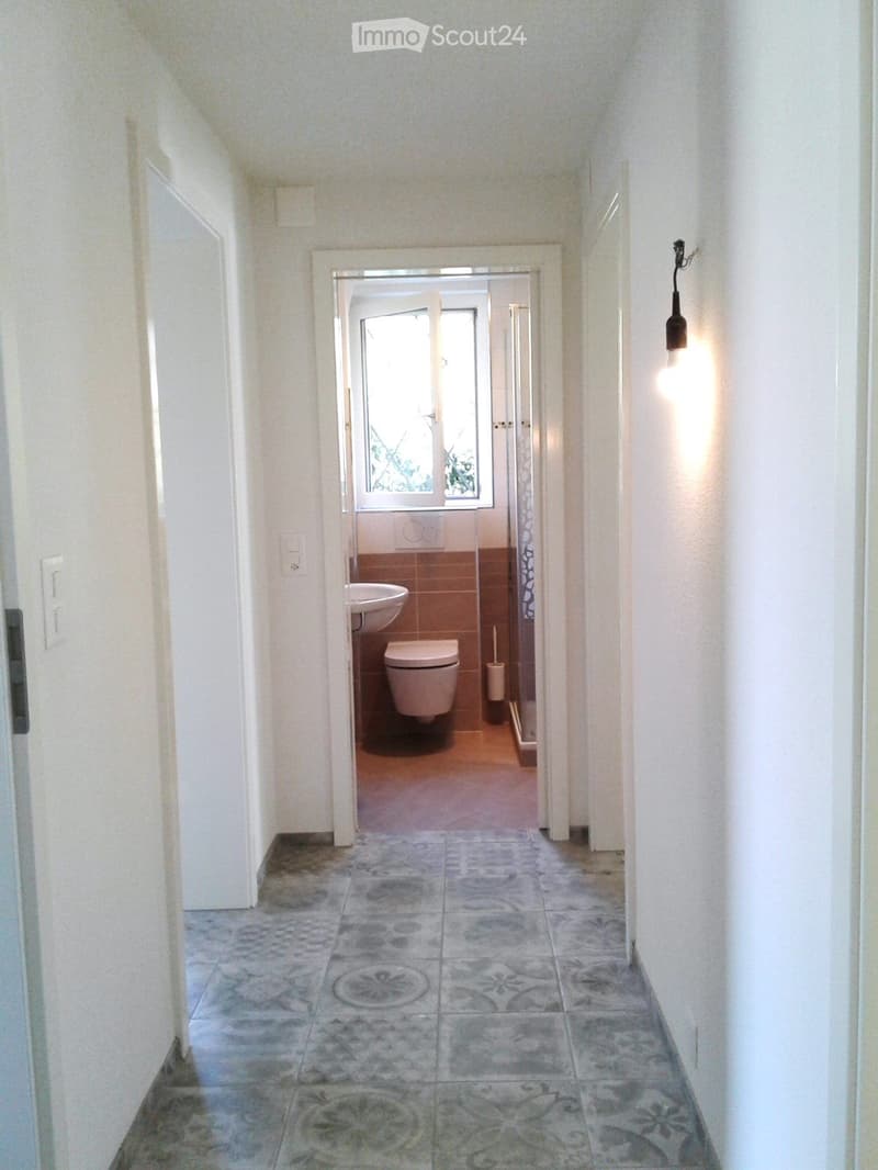 A narrow hallway with tiled flooring and white walls, leading to a small bathroom with a toilet, sink, and window.