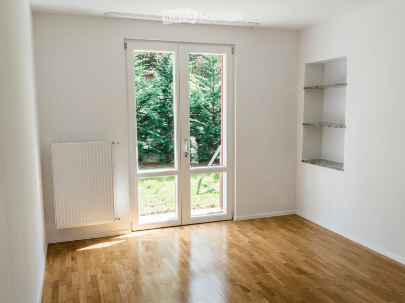 Empty room with light-colored walls, wooden flooring, and a large glass door leading to a garden area.