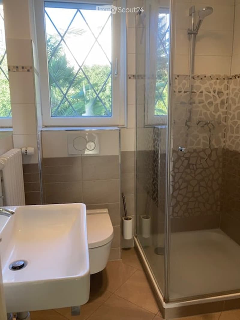 Modern bathroom with a glass-enclosed shower, a sink, a toilet, a radiator, and a window with decorative glass panes.