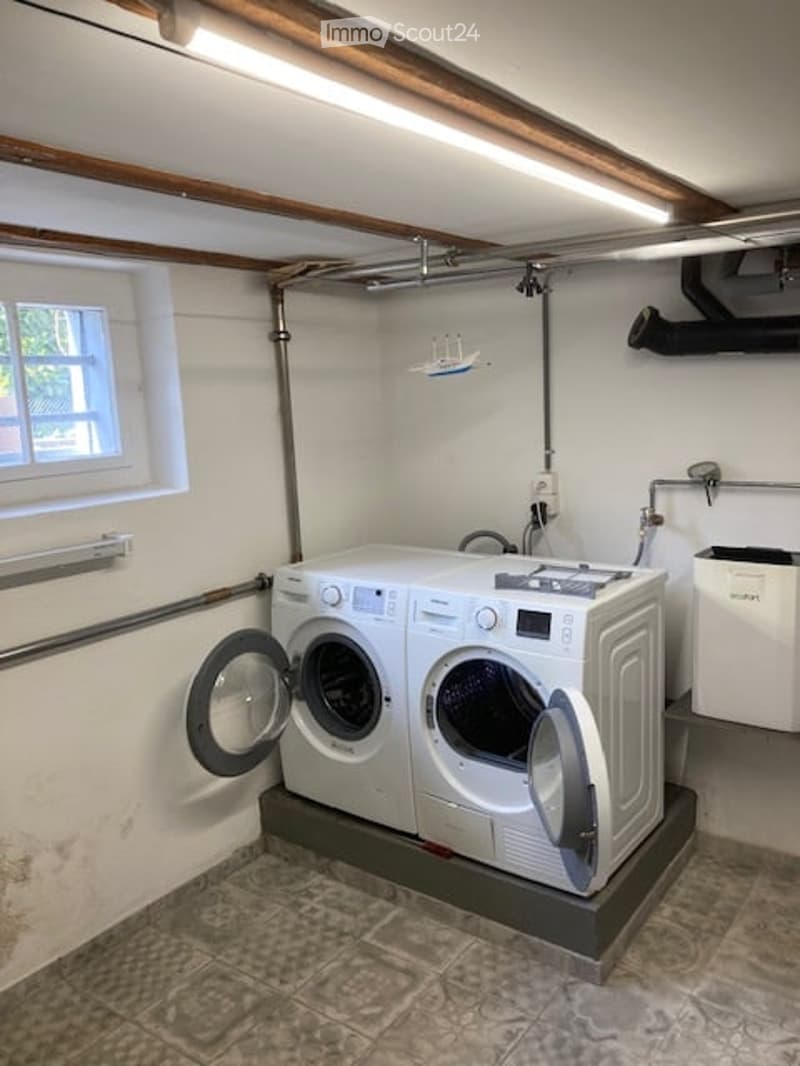 Laundry room with a washing machine and dryer, white walls, exposed wooden beams, and a window.