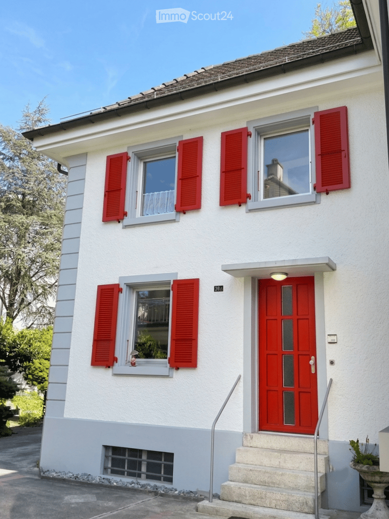 Two-story white house with red shutters and a red front door, featuring a small staircase leading to the entrance and a small garden area to the left.