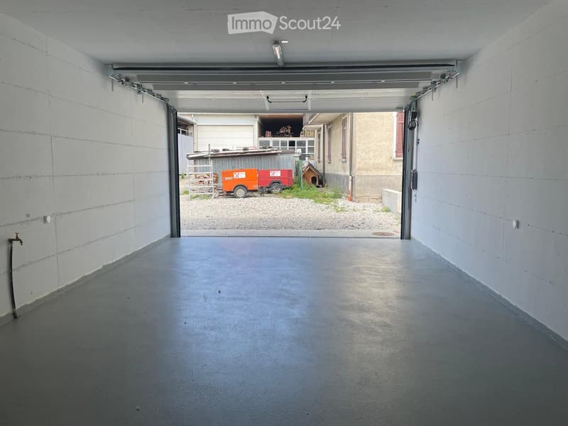 Empty garage with concrete floor, white walls, and an open door leading to an outdoor area with a trailer and small shed.