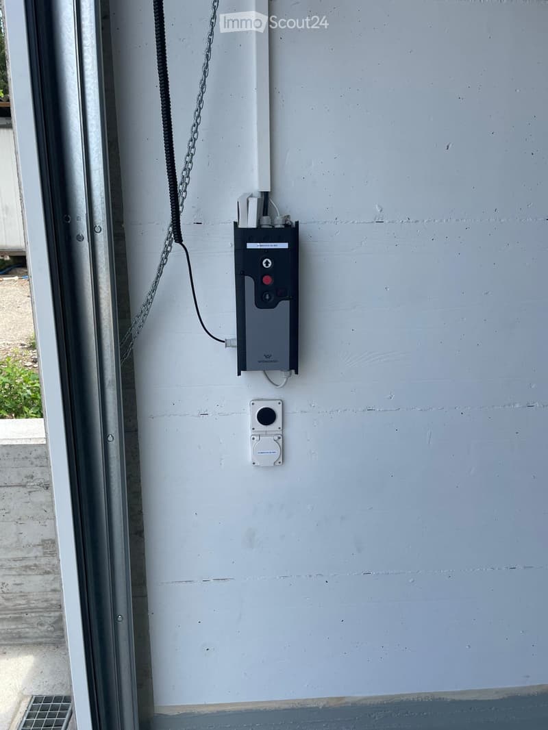 A close-up view of an electrical panel mounted on a light blue wall, featuring a black control unit with buttons and a digital display, along with a white switch and a black cable connected to the panel.