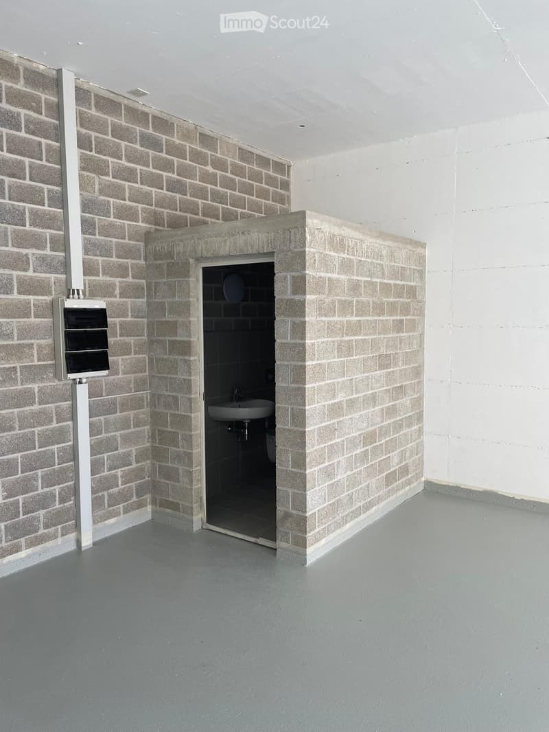 Empty room with grey brick walls and a grey concrete floor. A small, enclosed bathroom with a sink and toilet is visible through an open doorway.