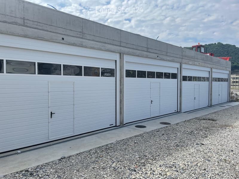 Four white garage doors with decorative windows, concrete roof, and gravel ground.