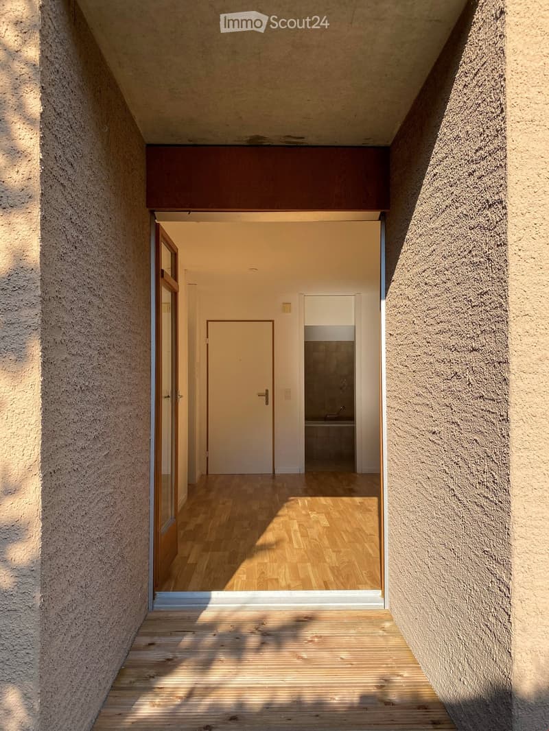 A narrow hallway with wooden flooring, textured beige walls, and a door leading to a room with a visible sink and tiled wall.