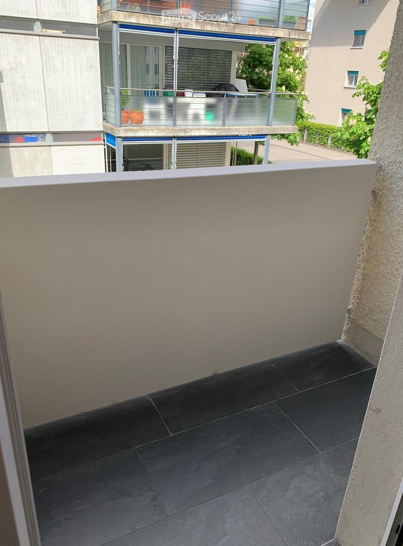 Balcony with dark tiled flooring, white railing, and visible neighboring building with balconies and potted plants.